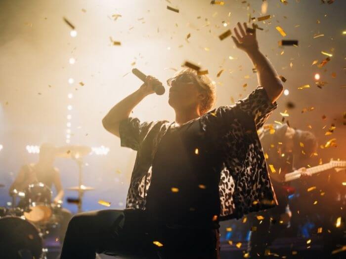Person performing on stage in a wheelchair, singing into a microphone with the band behind them, golden confetti streaming around them.