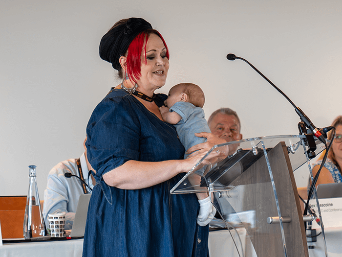 MU Executive Committe Vice Chair Andi Hopgood, stands and speaks from the mic while holding a small child.