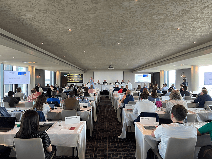Delegate conference attendees sit at tables in a bright room.