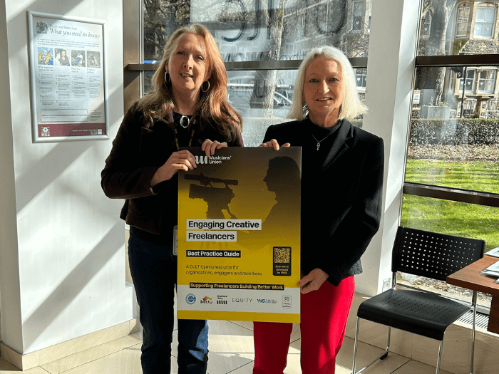 Ruth and Siân, standing in a bright indoor space holding a large poster of the front cover of the new Engaging Creative Freelancers Guide.