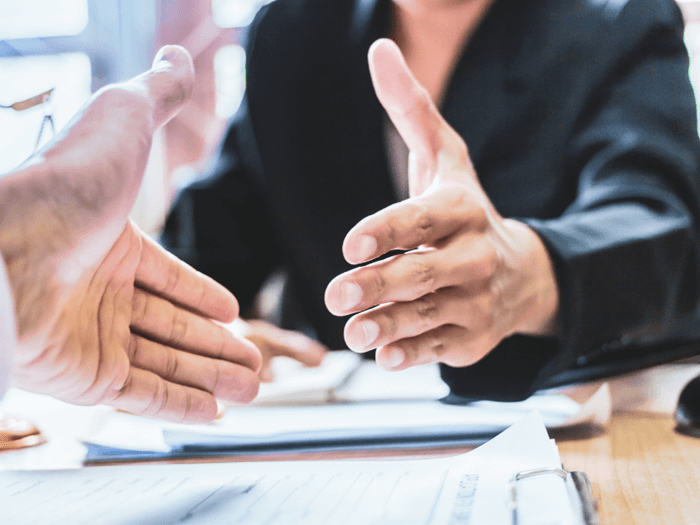 Two hands reaching across a desk with papers and clipboards to shake hands on an agreement.