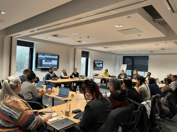 The MU Members' Assembly, sat in a semi-circle around the front desk and large monitor screens. Some people are making notes and others are typing on laptops.